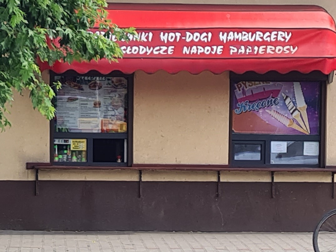 Storefront Zapiekanki Łask przy ulicy Warszawskiej 23. Czerwona markiza z napisem ZAPIEKANKI HOT-DOGI HAMBURGERY, dwa okna obsługi, drewniany blat dla klientów.