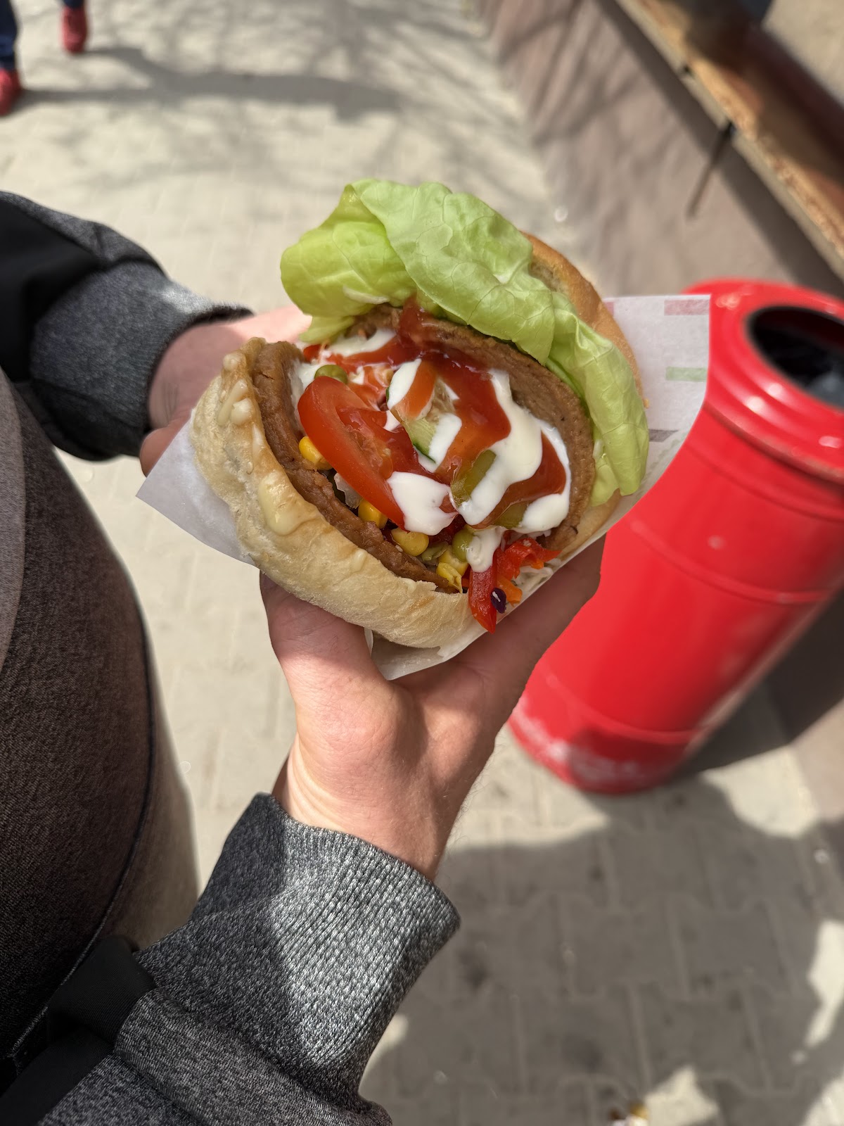 Hamburger w dłoni, z liściem sałaty, pomidorem, ketchupem i majonezem, w tle czerwony kosz na śmieci przy lokalu.